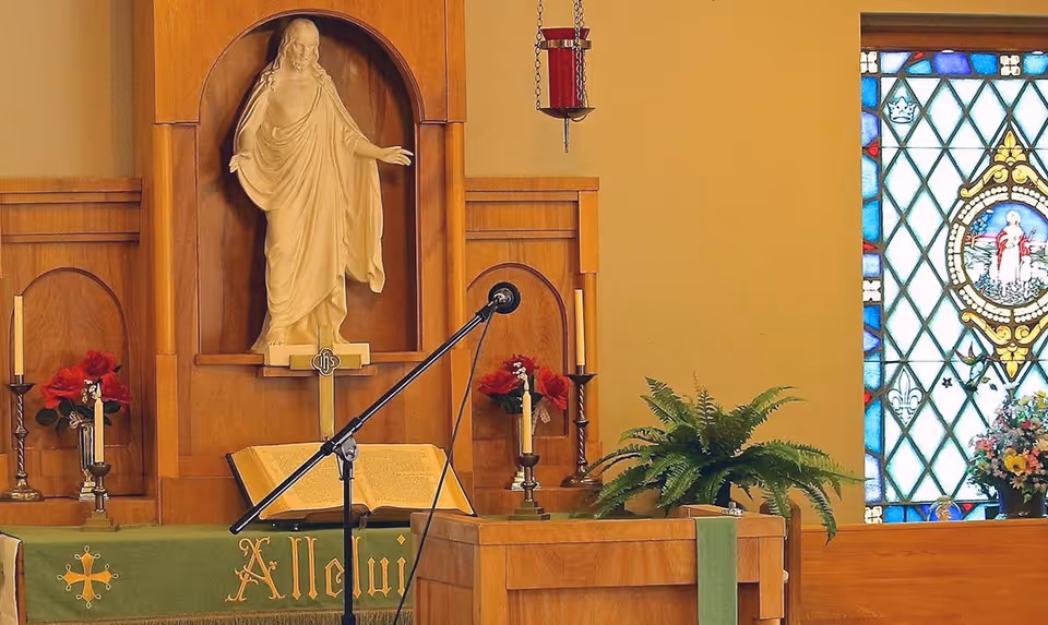 Interior view of a chapel area with a wooden altar featuring a statue of Jesus, an open Bible, candles, red flowers, a microphone stand, a green cloth with the word 'Alleluia', a hanging red candle holder, a green plant, and a stained glass window with religious imagery.