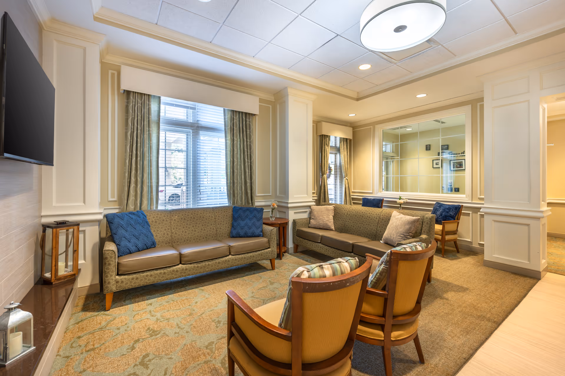 Bright, well-furnished senior living common room with sofas, armchairs, a wall-mounted TV, and a large window.