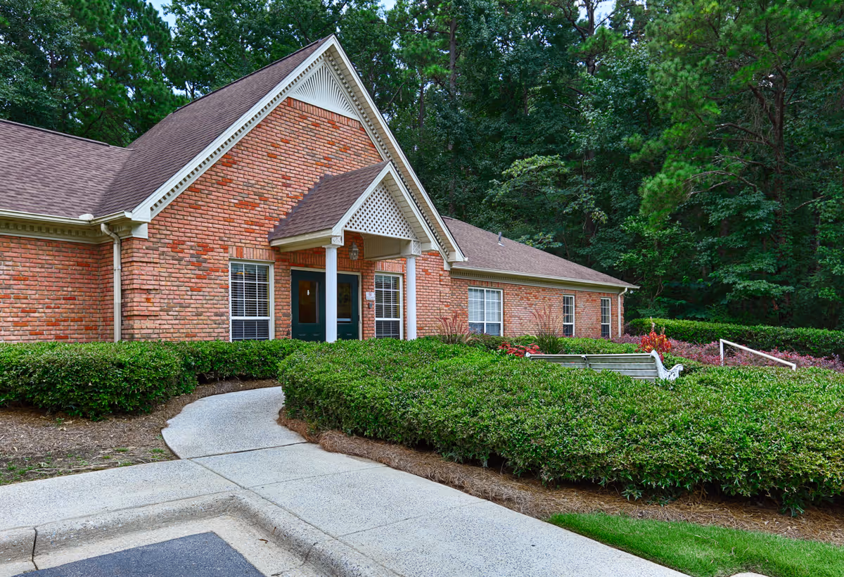 Exterior view of a single-story brick building with a peaked roof and a covered entrance supported by white columns. The building is surrounded by well-maintained green bushes and trees in the background. A concrete pathway leads to the entrance.