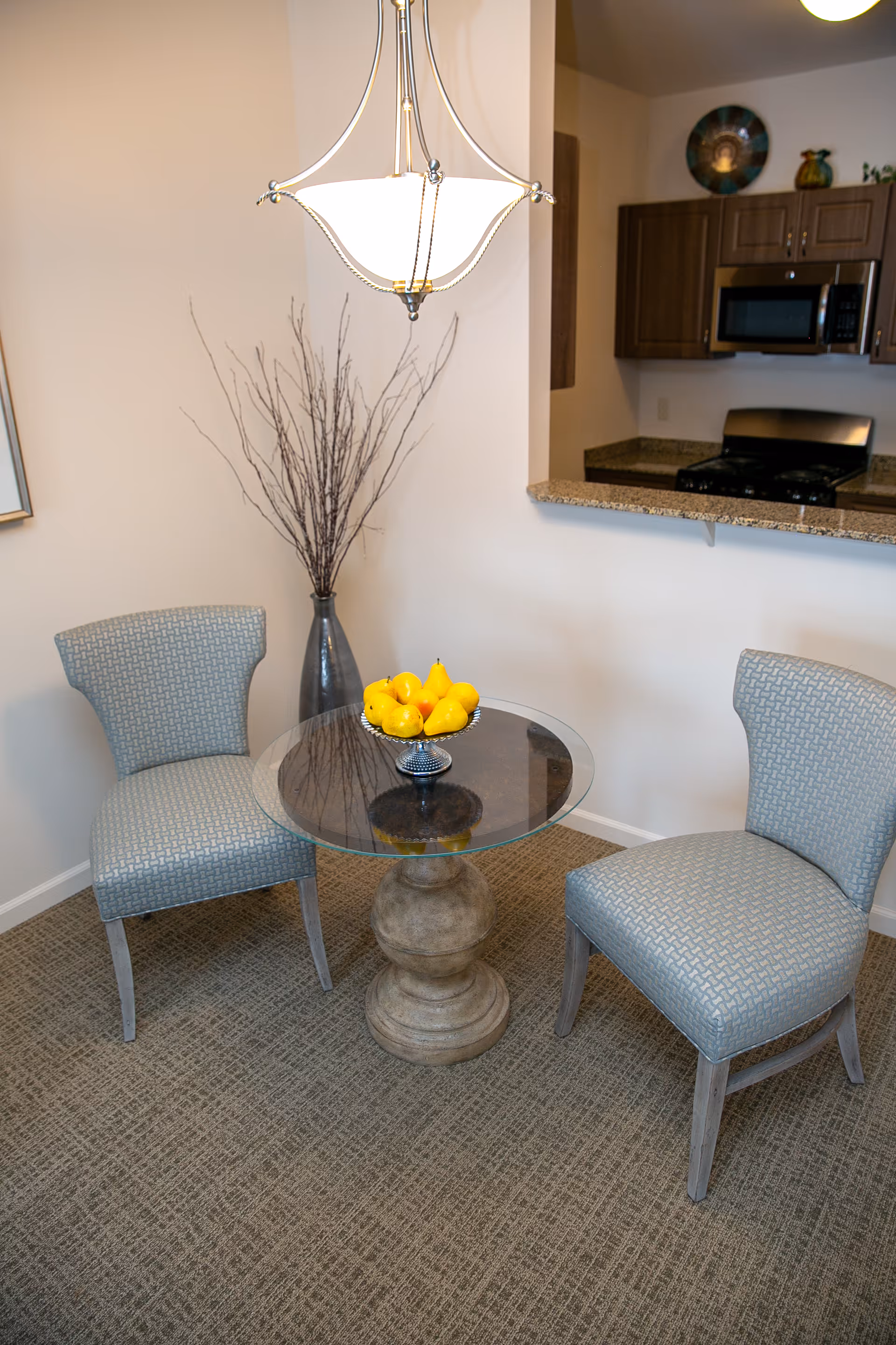 A small dining area with two upholstered chairs around a round glass-top table with a decorative pedestal base. A bowl of yellow pears is on the table. Behind the table is a tall vase with decorative branches. In the background, a kitchen area with wooden cabinets, a microwave, and a stove is visible.