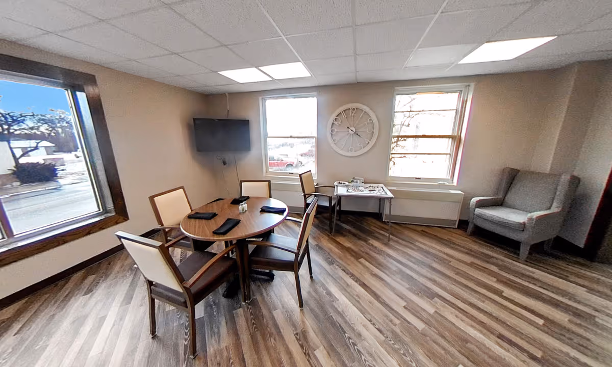 A small dining area with a round wooden table set with four chairs, each with a napkin on the table. The room has wood-patterned flooring, two large windows letting in natural light, a wall-mounted TV, a large round clock on the wall, a small table with items on it, and a gray armchair in the corner.