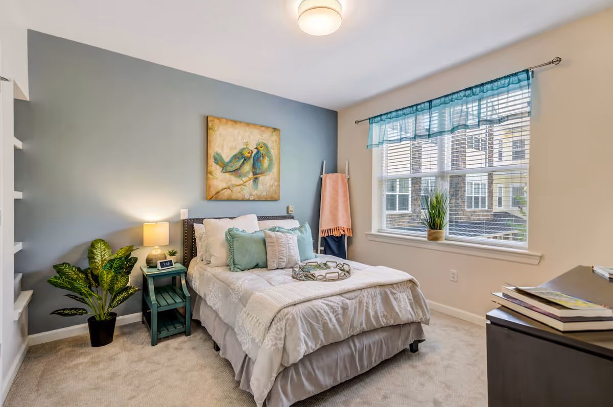 A cozy bedroom in an assisted living facility featuring a neatly made bed with white and teal pillows, a teal nightstand with a lamp and clock, a potted plant on the floor, a painting of two birds on the wall, a window with blinds and teal valance, and a dresser with books on top.