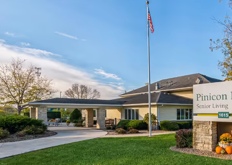 Front entrance of Pinicon Senior Living showing a covered driveway, flagpole, landscaped lawn, and facility sign.