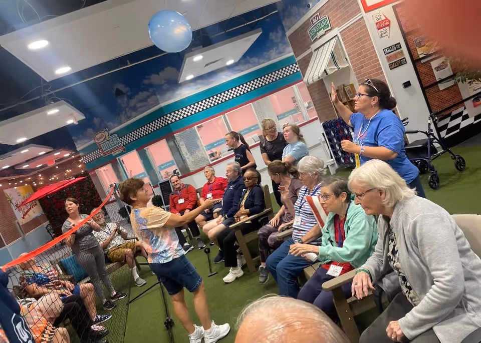 A group of elderly people and caregivers gathered indoors in a recreational area with artificial grass flooring. Some are seated on benches while others stand around a badminton net. The background features a colorful wall with a diner theme, including signs and a window with a striped awning. The atmosphere appears lively and social.