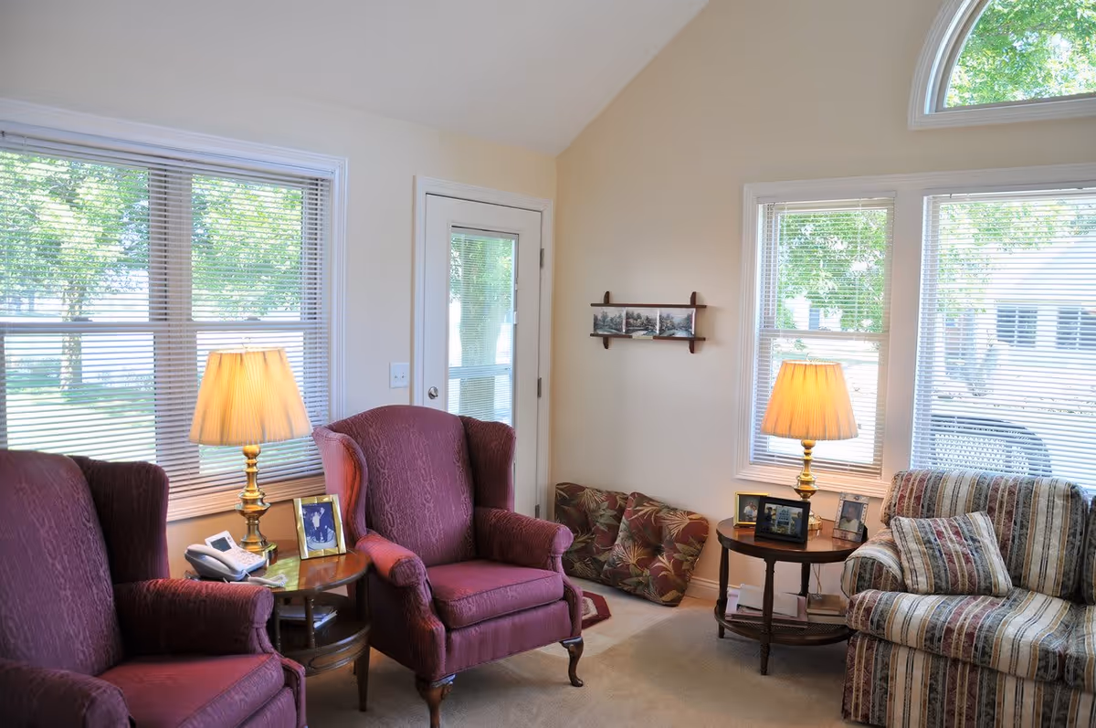 A cozy living room with two burgundy upholstered armchairs and a striped sofa. There are two wooden side tables with brass lamps and framed photos. Large windows with blinds let in natural light, and a door with a glass panel is visible. Cushions are placed on the floor against the wall.