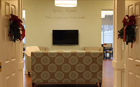 View through double doors decorated with red bows into a cozy living room area with a patterned sofa facing a wall-mounted flat screen TV. The wall above the TV has the text 'Grace House Where Family and Friends Gather'. A dining area with a table and chairs is visible in the background through an open doorway.