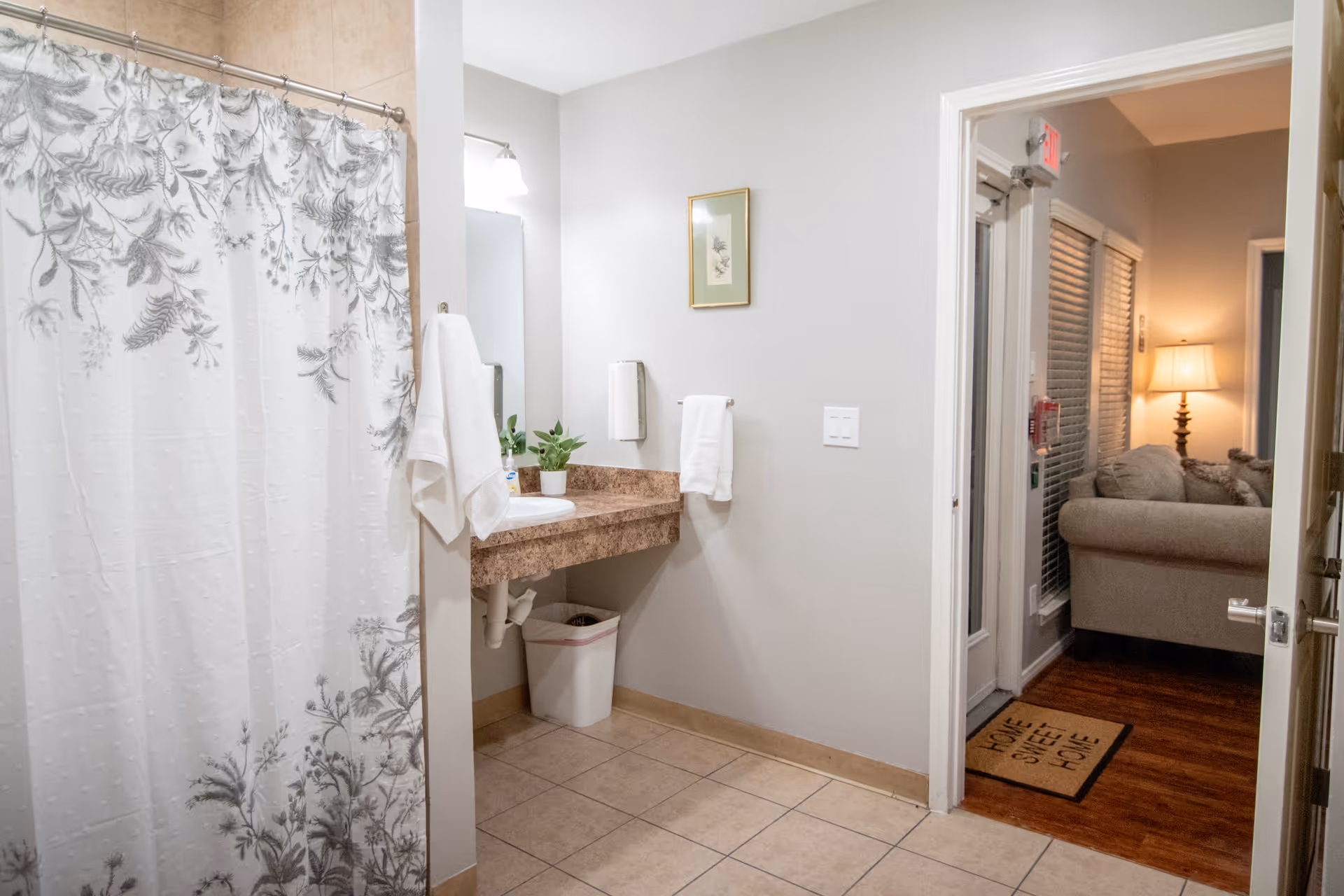 A bathroom with beige tiled floor and walls painted light gray. There is a shower with a white curtain featuring a gray botanical pattern on the left. A countertop with a sink, a small potted plant, and a soap dispenser is visible. Two white towels hang on the wall near the sink. The bathroom opens into a living room area with a beige couch, a lit lamp, and a 'Home Sweet Home' doormat on a wooden floor.