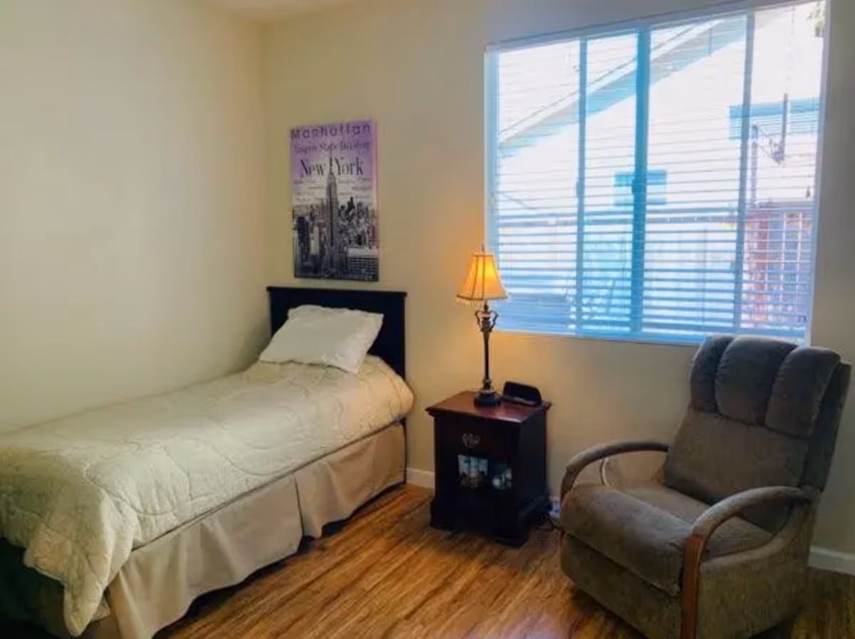 A small bedroom with a single bed covered with a beige quilt and a white pillow. Next to the bed is a wooden nightstand with a lamp and a small clock. A large window with blinds lets in natural light. There is a comfortable upholstered armchair with wooden armrests near the window. A framed picture of Manhattan, New York, hangs on the wall above the bed.