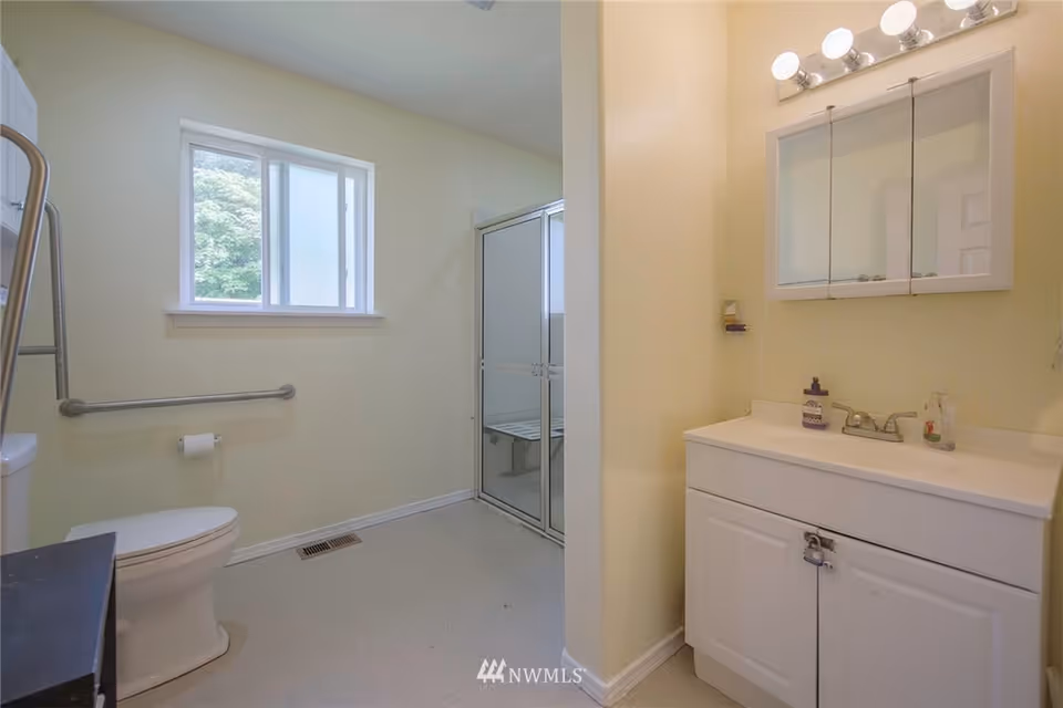 A clean bathroom with a white toilet, a grab bar on the wall, a window letting in natural light, a shower with sliding glass doors and a built-in bench, and a white vanity with a sink, faucet, and mirrored medicine cabinet above it.