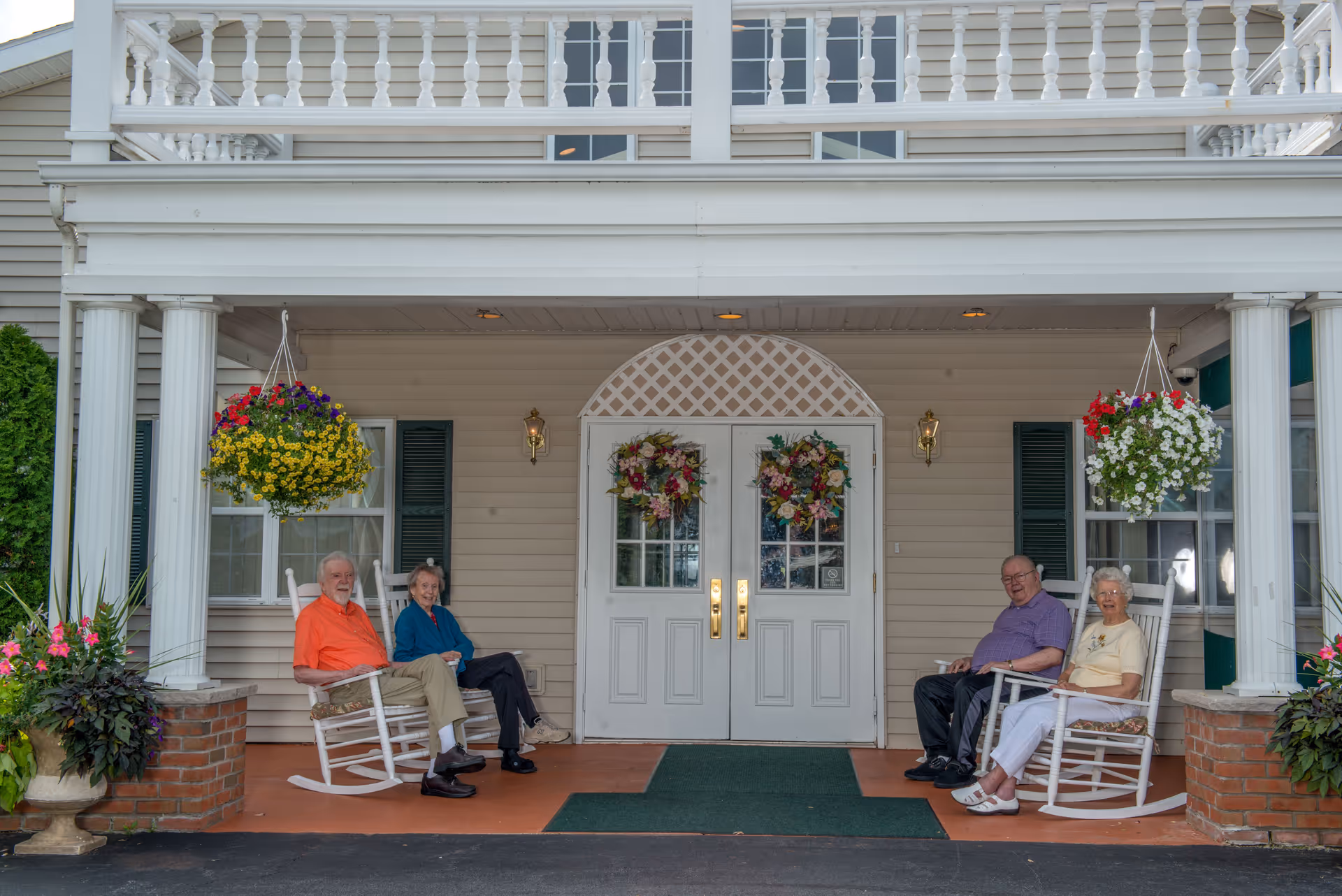 Four elderly people sitting on white rocking chairs on the covered porch of a building with white double doors decorated with floral wreaths. There are hanging flower baskets and potted plants on either side of the porch.