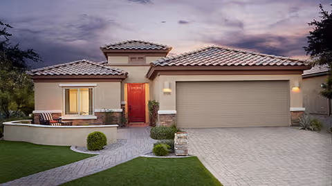 Front view of a single-story stucco house with a tiled roof, red front door, two-car garage, driveway, and landscaped lawn at dusk.