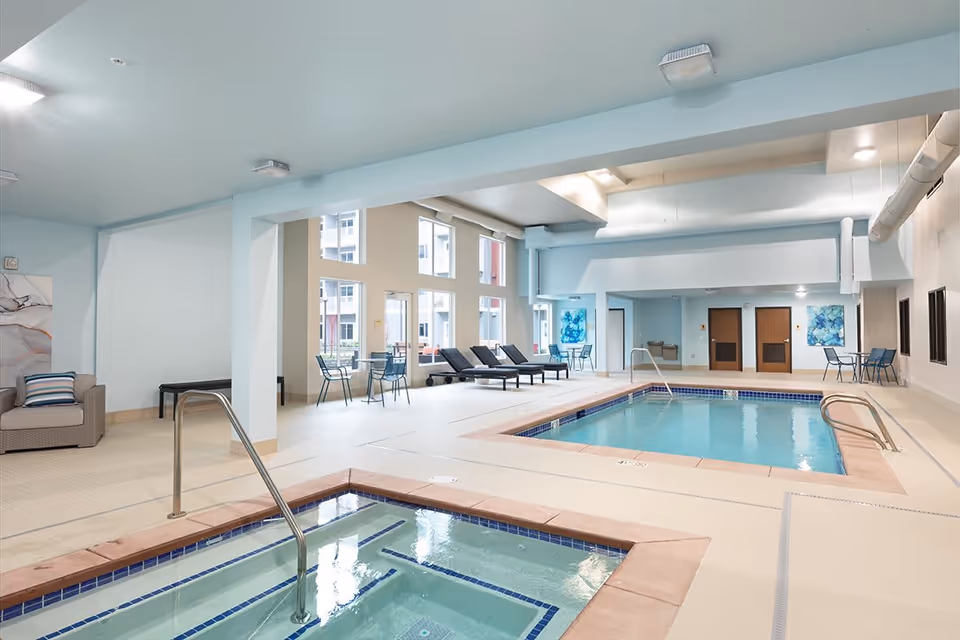 Indoor pool and hot tub area in a senior living facility with lounge chairs, tables and large windows.