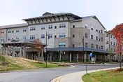 Three-story senior living building with a covered main entrance, stone and siding exterior, driveway and landscaped lawn.
