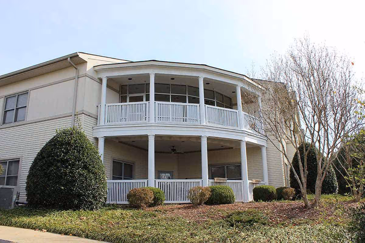 Exterior view of a two-story senior living community building with a covered balcony on the upper floor and a covered porch on the lower floor, surrounded by bushes and trees with a clear sky in the background.