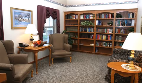 A cozy interior room with two upholstered armchairs and a wooden side table with a lamp and a telephone. There is a large wooden bookshelf filled with books and decorative plants against the wall. Another side table with a lamp is next to a patterned sofa. The room has carpeted flooring, a window with burgundy curtains, and a framed painting on the wall.