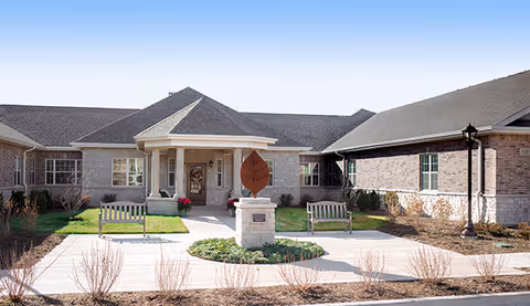 Front exterior view of Franklin Place Memory Care facility with a covered entrance, two benches, a decorative sculpture on a pedestal, and landscaped areas with grass and shrubs.