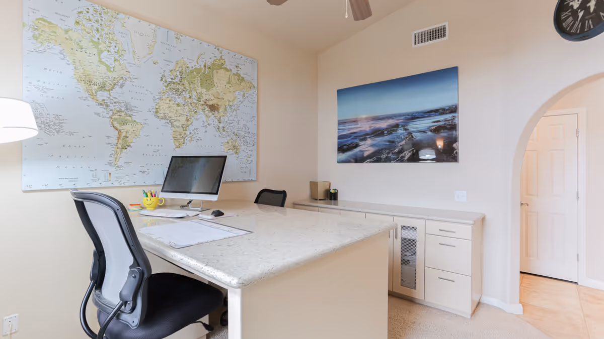 A bright office space with a large world map on the wall, a desk with a computer monitor, office chair, and various stationery items. There is a scenic photograph of a rocky shoreline on another wall, a clock above an arched doorway, and built-in cabinets beneath the photograph.