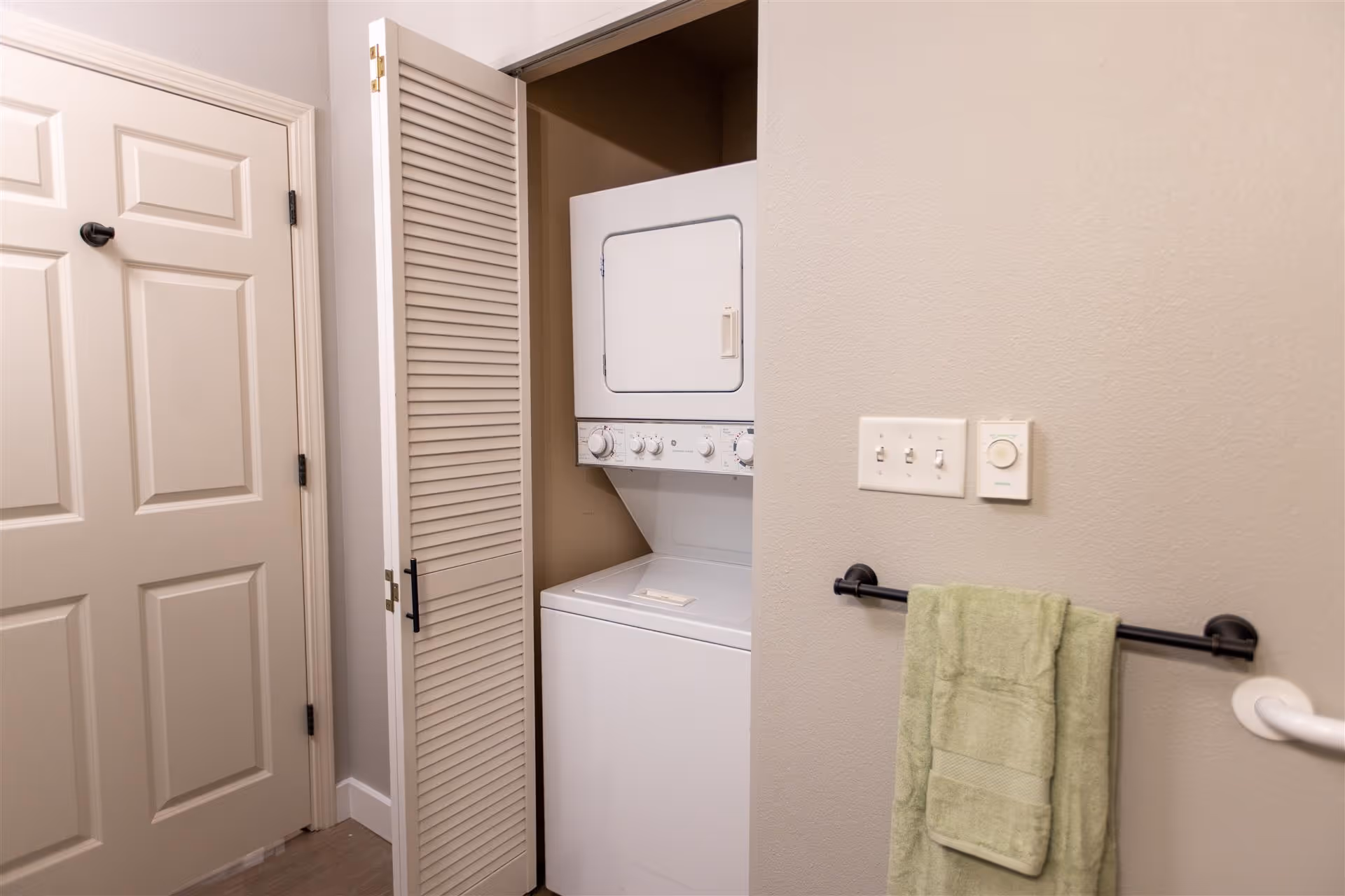 A small laundry area with a stacked washer and dryer unit inside a closet with louvered doors, adjacent to a beige wall with a towel rack holding a green towel and light switches above it.