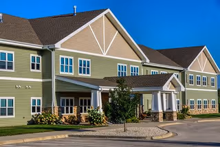 Two-story green and beige building with a covered main entrance, landscaping, and a circular driveway under a clear blue sky.