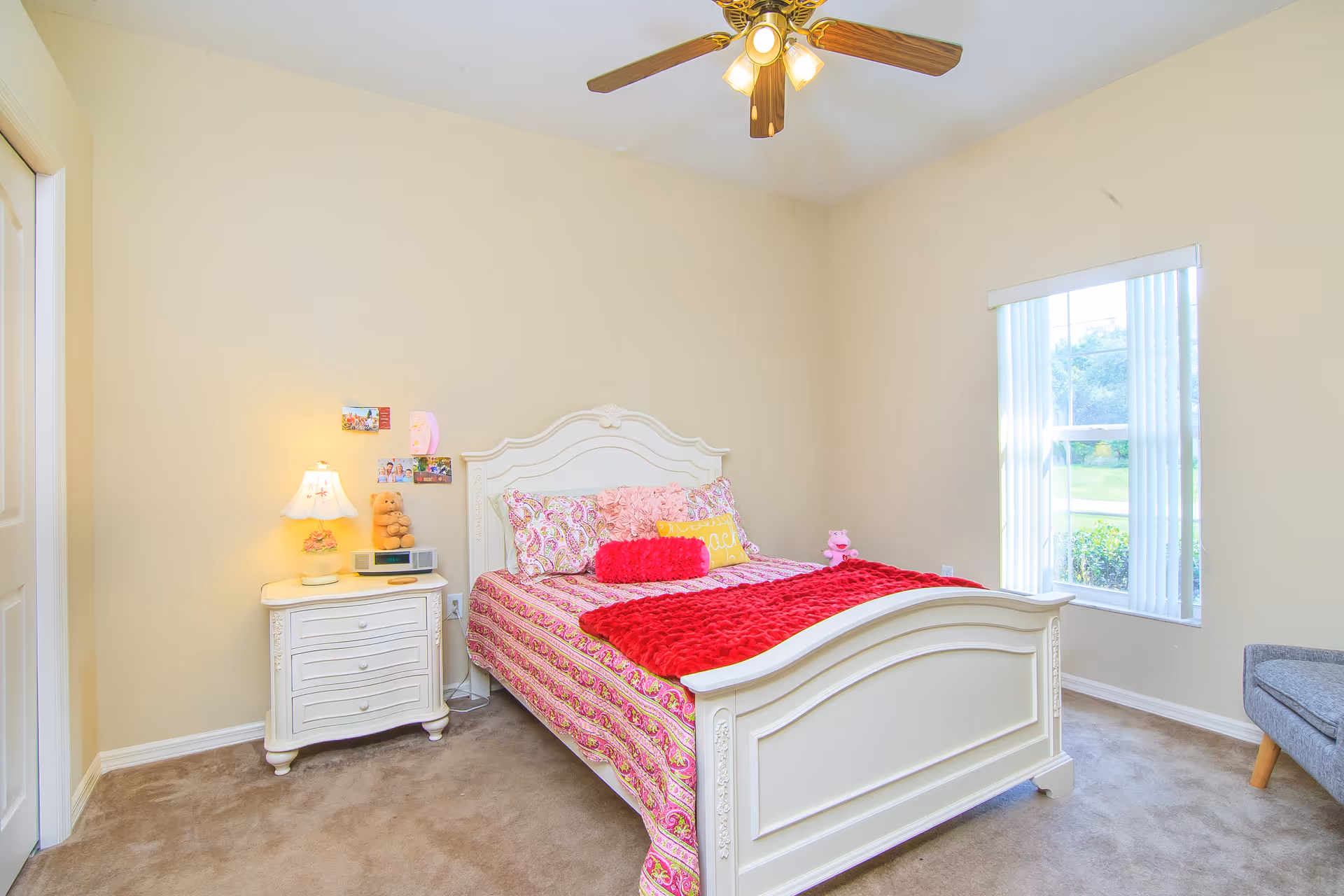 A bright bedroom with a white wooden bed frame and a colorful pink and red bedspread. There is a white nightstand with a lamp, a clock, and a teddy bear on it. A window with vertical blinds lets in natural light, and a ceiling fan with lights is mounted on the ceiling. The room has beige walls and carpeted flooring.