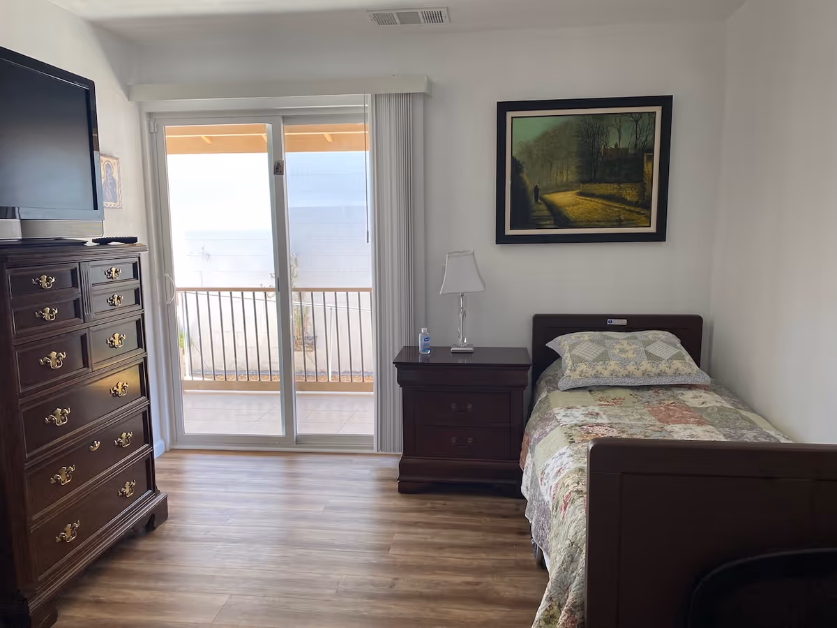 A small bedroom with a single bed covered in a patchwork quilt, a wooden nightstand with a lamp and a bottle of hand sanitizer, a wooden dresser with a flat-screen TV on top, and a sliding glass door leading to an outdoor patio area. A framed painting hangs on the wall above the bed.