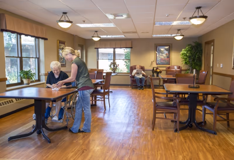 A bright communal dining/activity room with tables and chairs, a caregiver assisting an elderly woman in a wheelchair and another resident seated in the back reading.