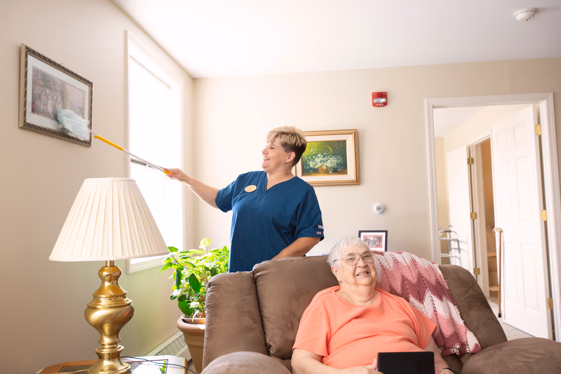A caregiver in blue scrubs dusts a framed picture on the wall near a window in a cozy living room while an elderly woman with glasses and a peach-colored shirt sits comfortably on a brown recliner with a pink and white crocheted blanket draped over the back, smiling and holding a tablet.