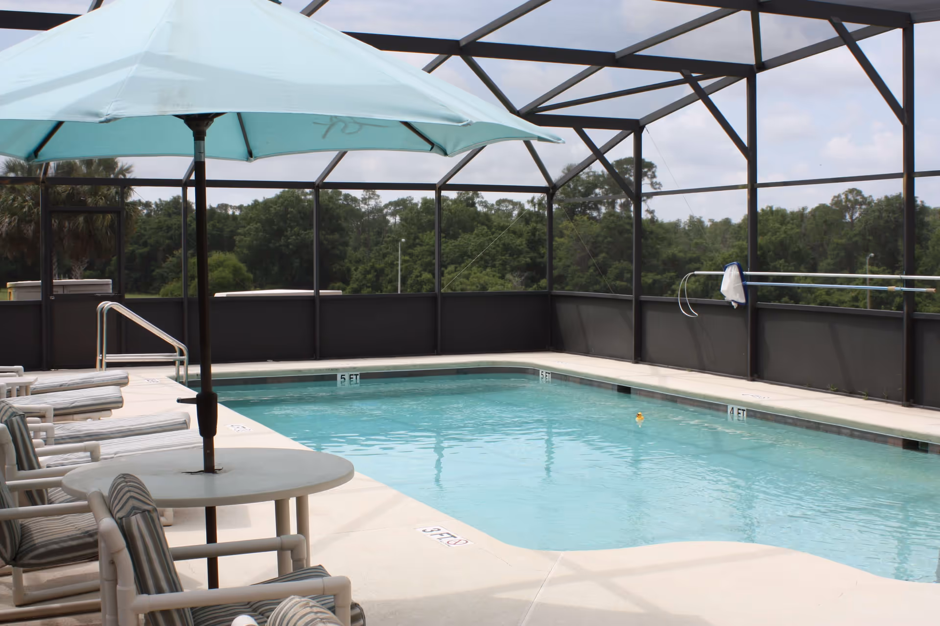 Outdoor swimming pool area enclosed by a screened structure with a light blue umbrella shading a round table surrounded by cushioned chairs. The pool has depth markers and a pool cleaning net is hanging on the enclosure. Trees and a partly cloudy sky are visible in the background.