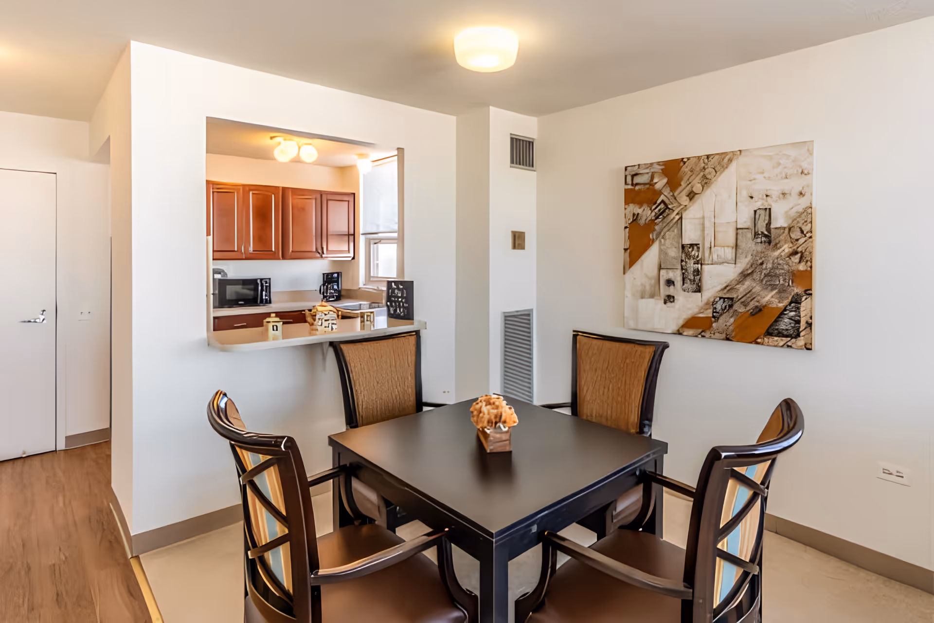 A dining area with a square black table surrounded by four chairs with brown cushions. On the table is a small decorative item. The dining area is adjacent to a kitchen with wooden cabinets, a microwave, and a coffee maker visible through a pass-through window. A modern abstract painting hangs on the white wall to the right.