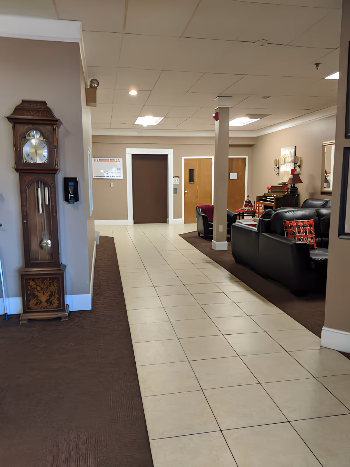 Interior hallway of a senior living facility with beige tiled floor and brown carpet on the sides. On the left side, there is a tall wooden grandfather clock and a wall-mounted hand sanitizer dispenser. On the right side, there is a seating area with black leather sofas, a small table with Christmas decorations, a piano, and wall art. The hallway leads to three closed doors at the end.