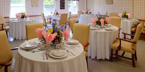 Round tables with white linens, floral centerpieces, and yellow-upholstered chairs in a dining room.