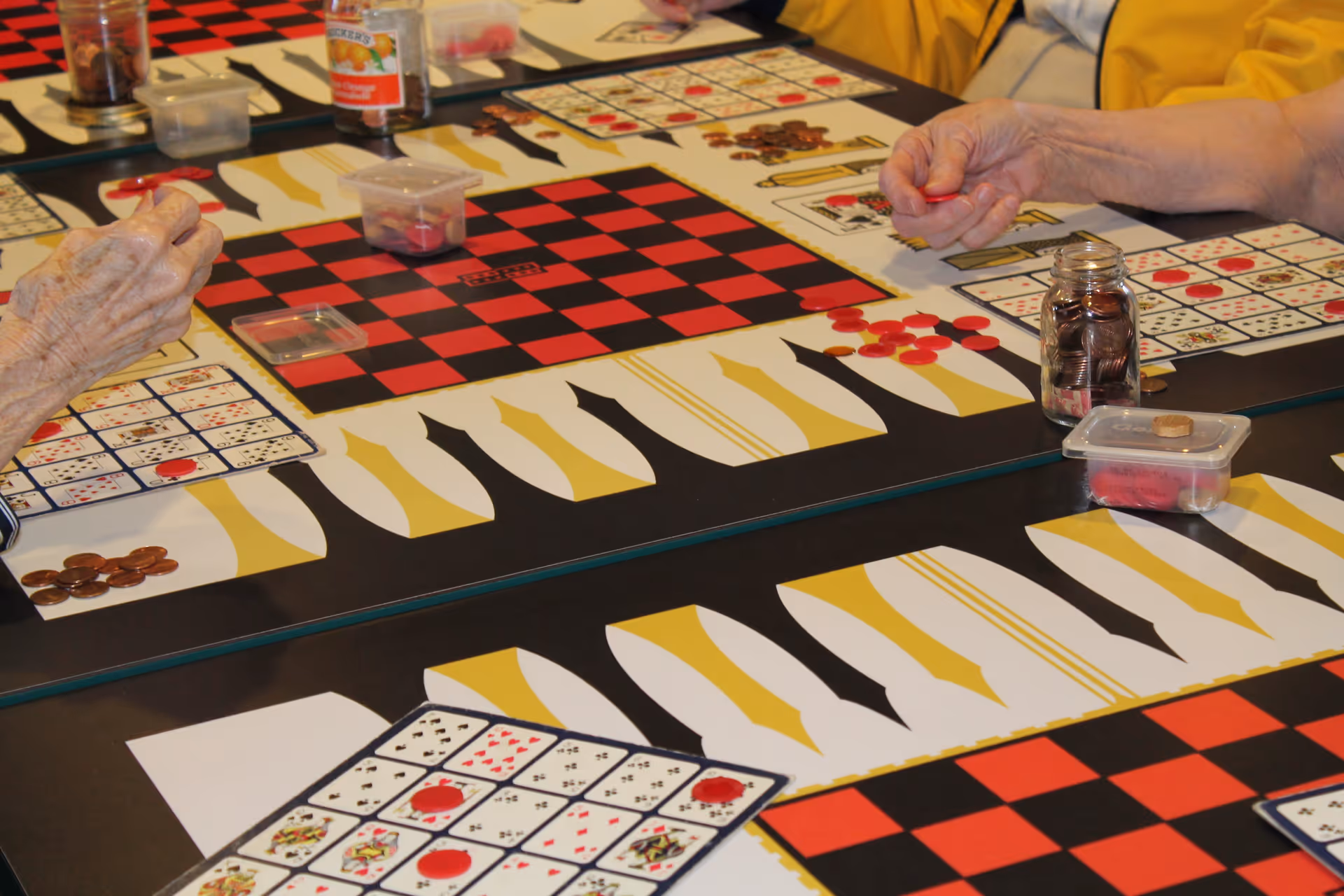Close-up of elderly hands playing a board game with red and black checkered patterns and cards, with game pieces and coins on the table.