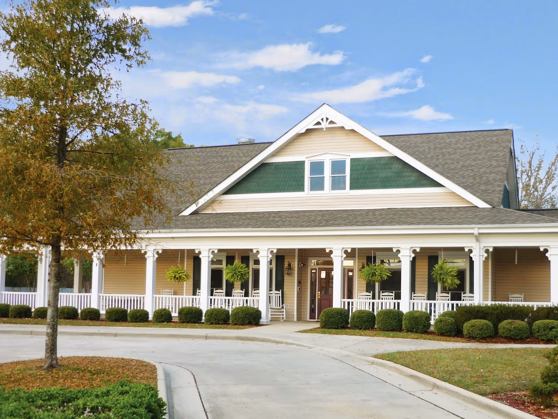 Front exterior view of a single-story building with a large covered porch featuring white railings and rocking chairs. The building has beige siding with green accents and a gray shingled roof. There are hanging plants on the porch and neatly trimmed bushes along the front. A tree and landscaped areas are visible in the foreground under a partly cloudy sky.