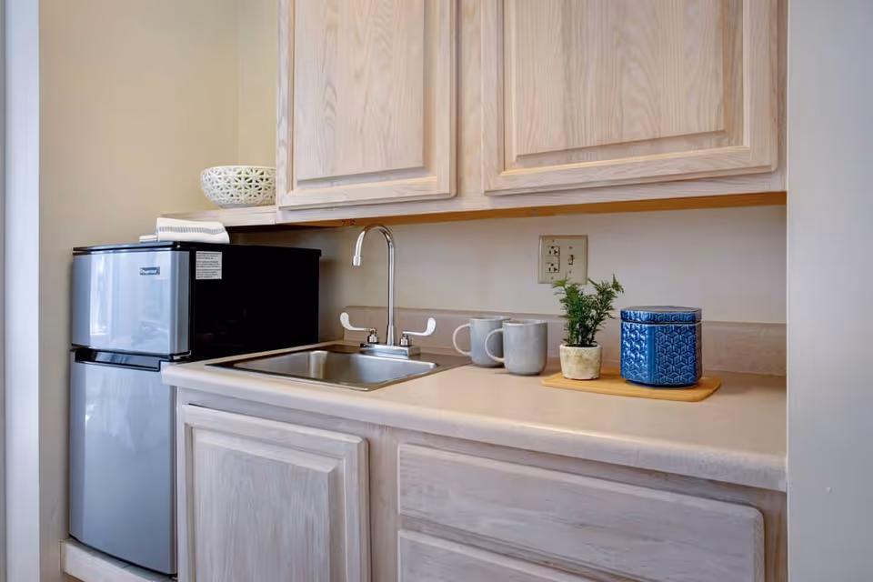 A small kitchenette area with light wood cabinets, a stainless steel sink with a faucet, a mini refrigerator, a microwave, two gray mugs, a small potted plant, and a blue decorative container on a beige countertop.