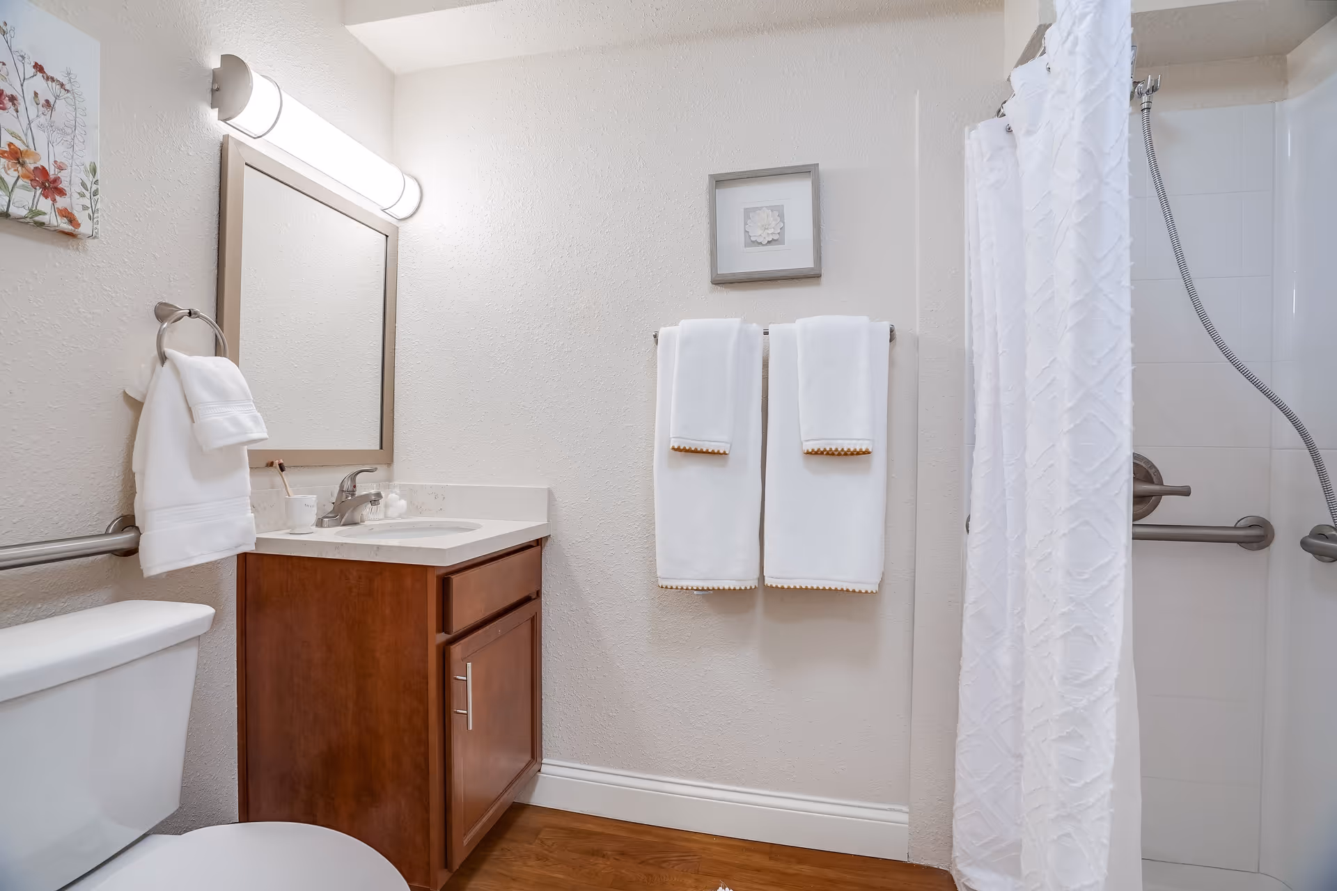 A clean and well-lit bathroom with a white toilet, wooden vanity with a sink, a large mirror above the sink, two white towels hanging on a towel rack, a white shower curtain, and grab bars in the shower area. The walls are light-colored and there is a small framed picture above the towels.