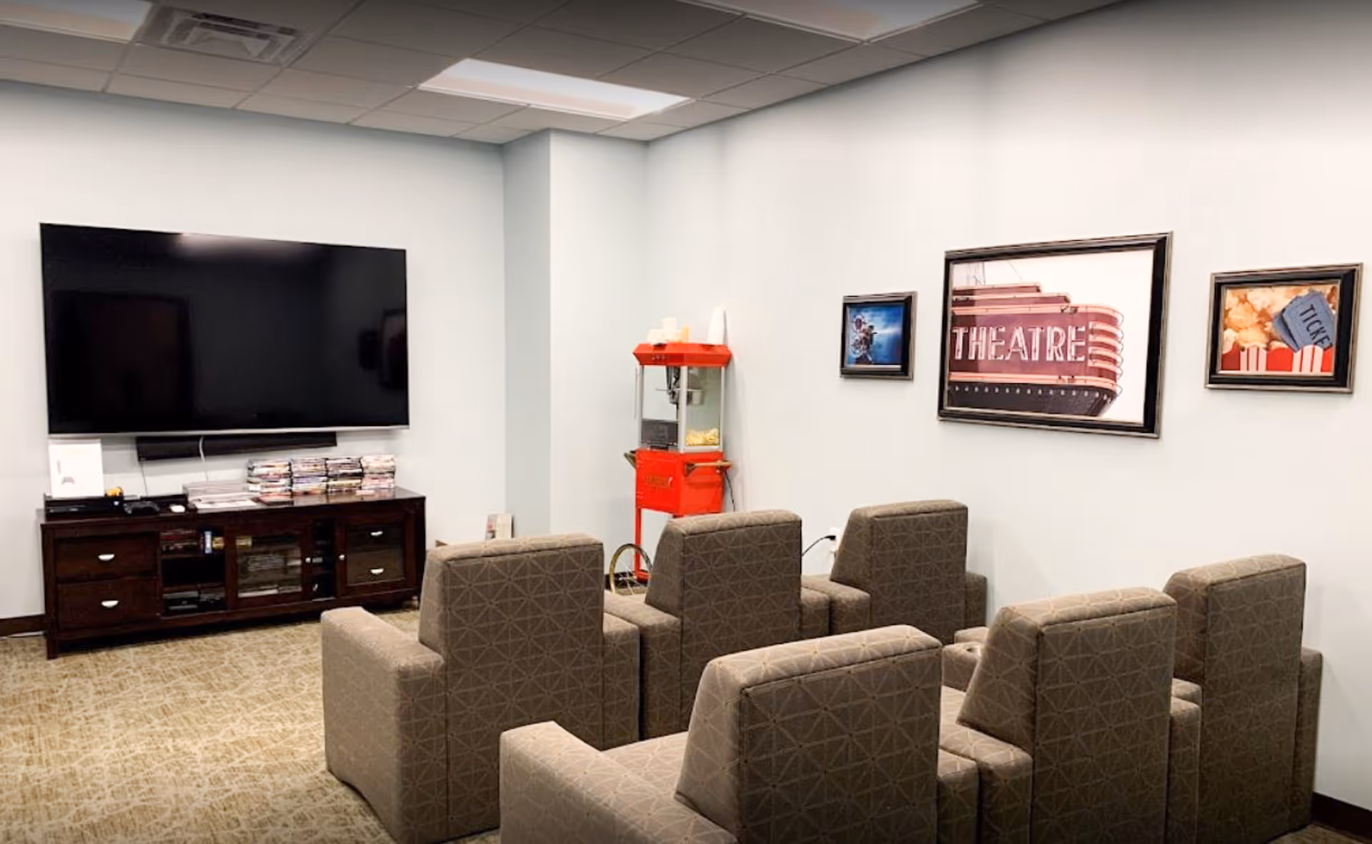 A small movie theater room with six cushioned armchairs arranged in two rows facing a large flat-screen TV mounted on the wall. Below the TV is a dark wooden cabinet with DVDs and other media items. A red popcorn machine is positioned in the corner, and three framed pictures related to movies and theater hang on the adjacent wall.