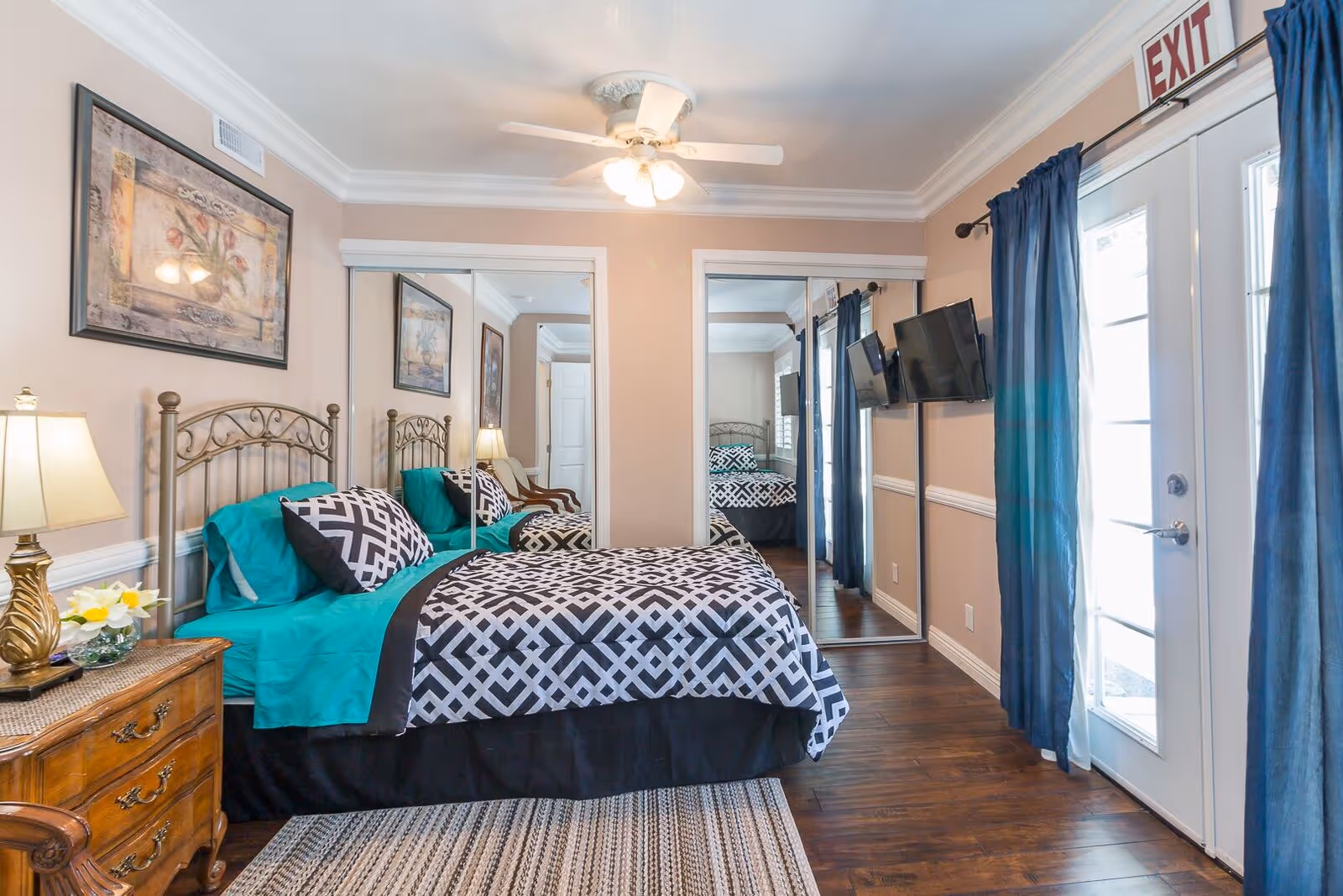 A cozy bedroom with a double bed featuring teal and black patterned bedding. The room has wooden flooring, a ceiling fan with lights, and mirrored sliding closet doors. There is a wooden nightstand with a lamp and flowers, a wall-mounted TV, blue curtains on a glass door, and framed artwork on the wall.