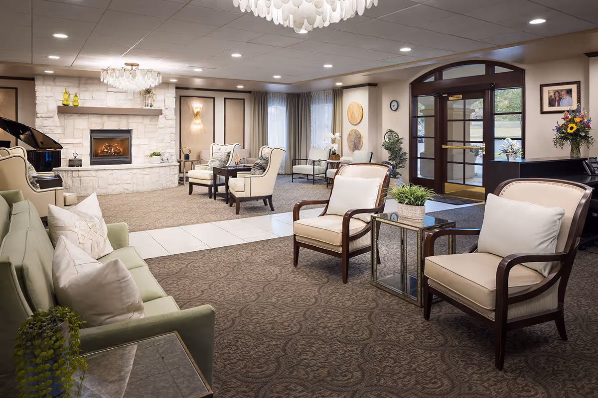 A spacious and well-lit senior living facility common area with comfortable seating including armchairs and a green sofa. The room features a stone fireplace with a fire burning, a grand piano, decorative wall art, plants, and a glass door entrance. The carpeted floor and soft lighting create a warm and inviting atmosphere.