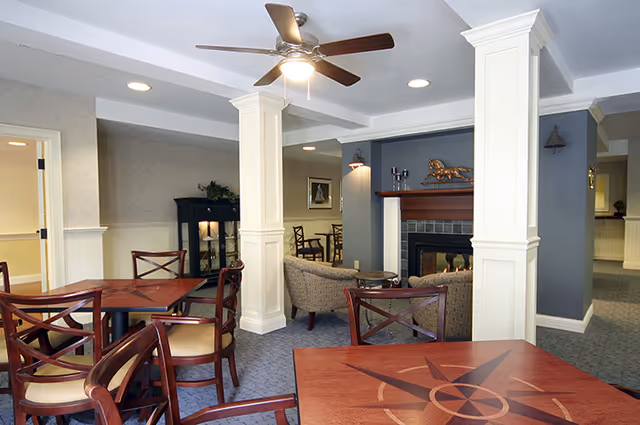 A cozy senior living common area with wooden tables and chairs, a ceiling fan with light, a fireplace with decorative items on the mantel, and comfortable armchairs arranged around the fireplace. The room features white pillars, soft lighting, and carpeted floors.