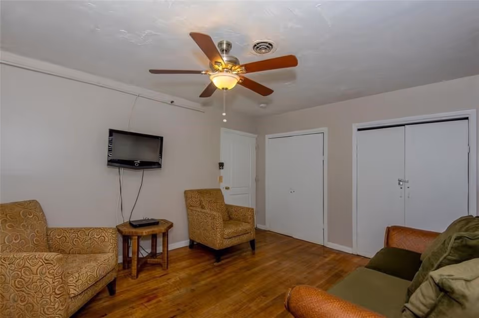 Small living room with armchairs, a sofa, a wall-mounted TV, a ceiling fan, and hardwood floors.