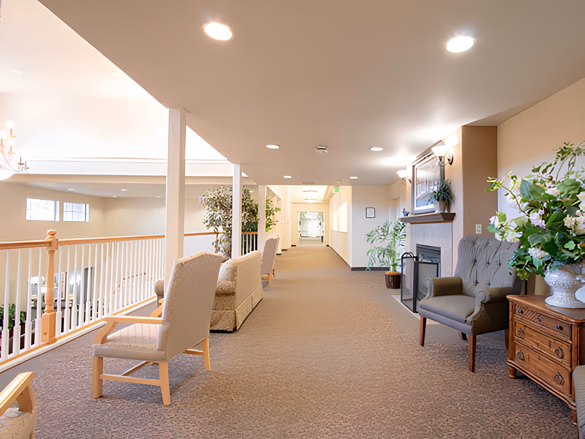 A bright and spacious senior living facility hallway with beige walls and carpeted floor. The hallway features several upholstered chairs and sofas arranged along the sides, potted plants, a fireplace with a framed picture above it, and soft ceiling lights. A white railing runs along the left side overlooking a lower level.