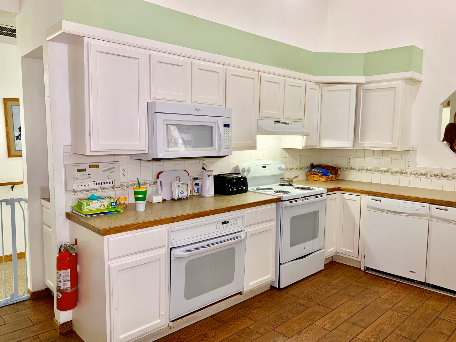 A clean kitchen with white cabinets and appliances including a microwave, oven, stove, and dishwasher. The countertop has various items such as a toaster, a basket, a Subway cup, and some kitchen essentials. The floor is wooden, and there is a fire extinguisher mounted on the side of the counter.