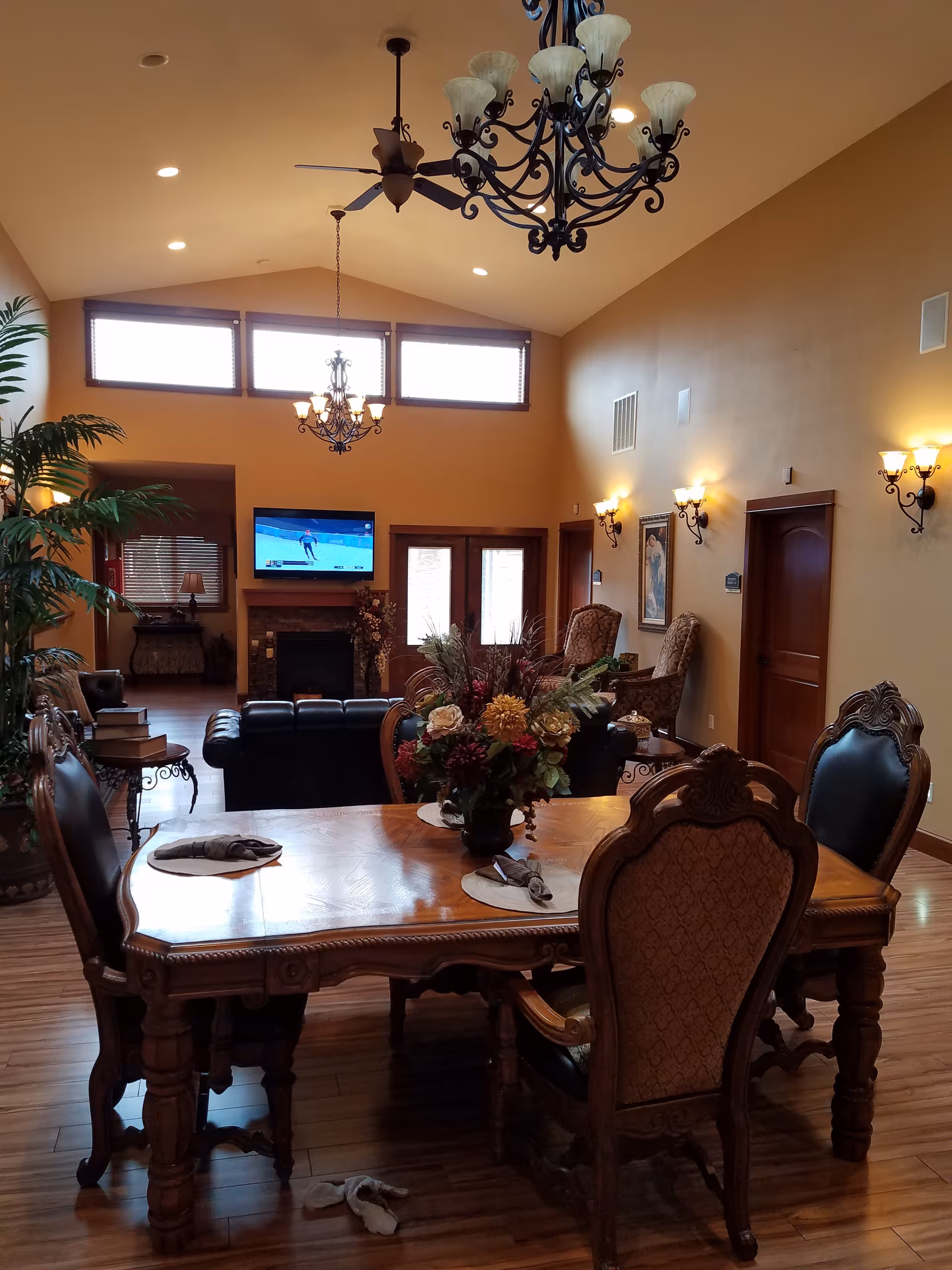 A warmly lit living room and dining area with a wooden dining table set with four ornate chairs and a floral centerpiece. In the background, there is a fireplace with a TV mounted above it, a black leather couch, two armchairs, wall sconces, and large windows near the ceiling letting in natural light.