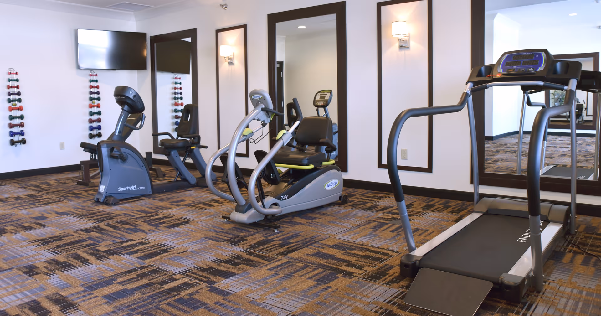 A fitness room with exercise equipment including a treadmill, a recumbent bike, and an elliptical machine. There are wall-mounted racks holding colorful dumbbells and large mirrors on the walls. The room has patterned carpet flooring and white walls with dark trim.
