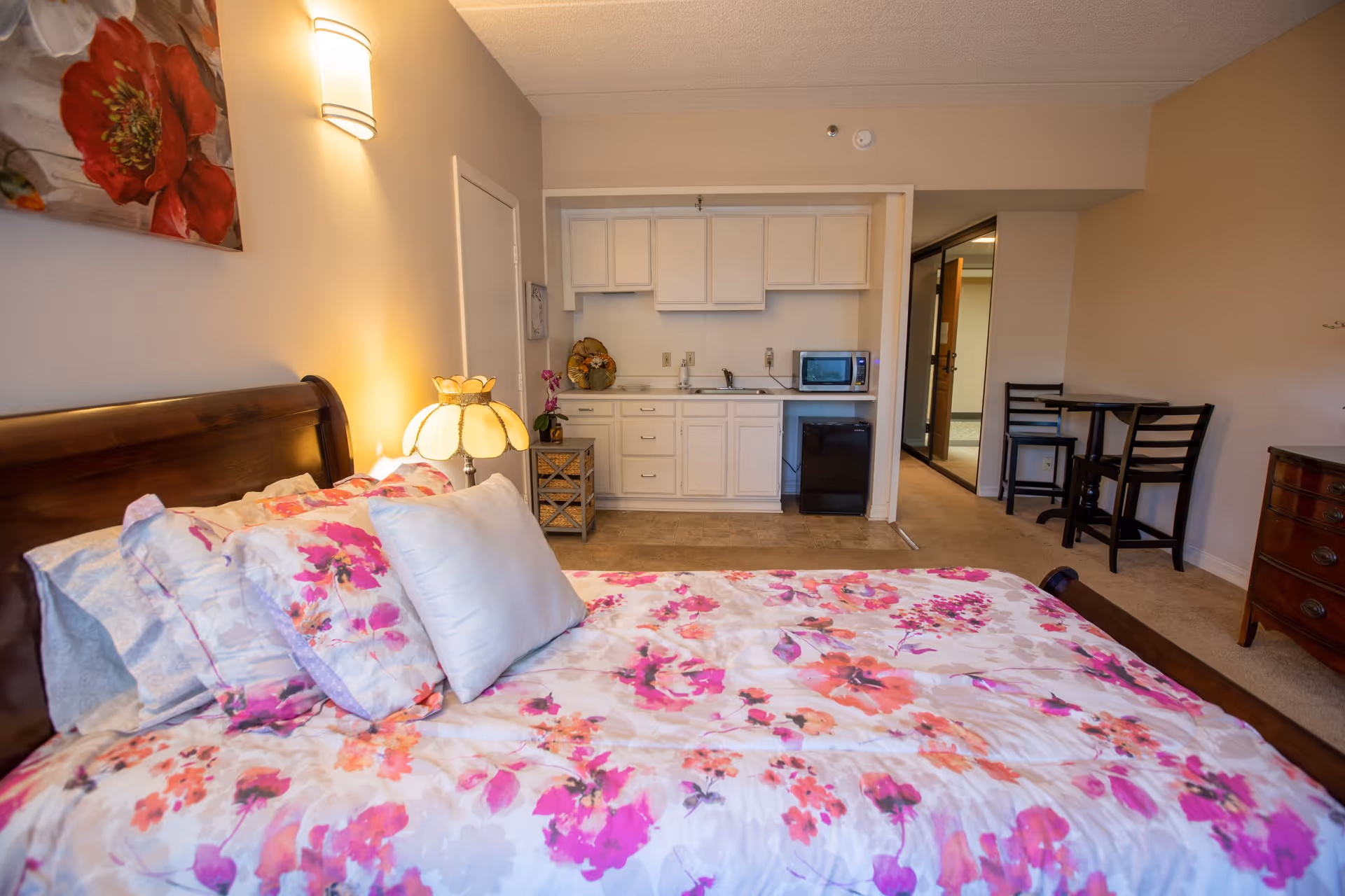 A cozy bedroom with a wooden bed frame and floral bedding in shades of pink, purple, and orange. Next to the bed is a small table with a decorative lamp and a wall-mounted light fixture above. In the background, there is a small kitchenette with white cabinets, a microwave, a mini fridge, and a small dining table with two chairs. The walls are painted beige and the room has a warm, inviting atmosphere.