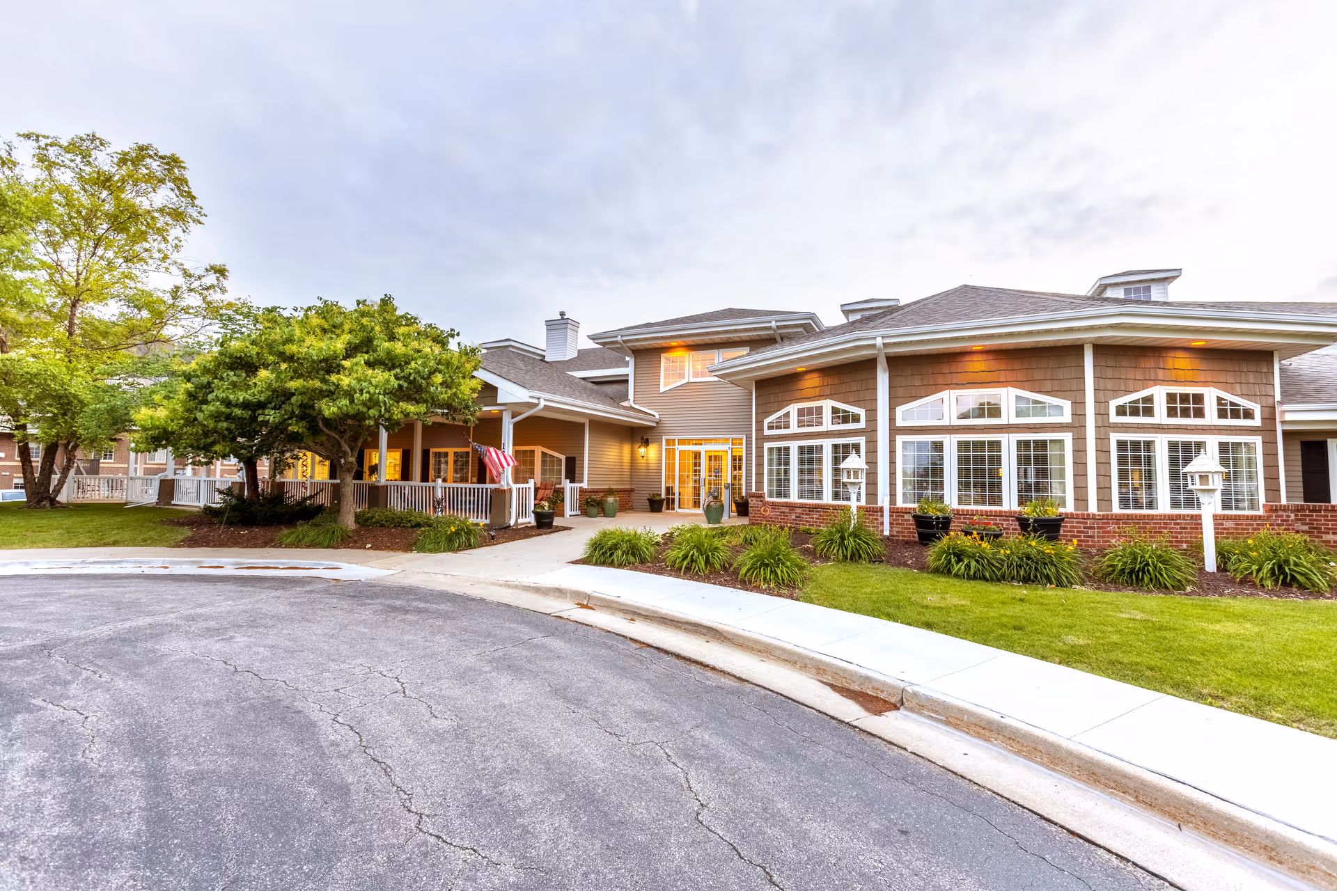 Exterior front entrance of a senior living facility with a covered porch, large arched windows, landscaping, and a curved driveway.