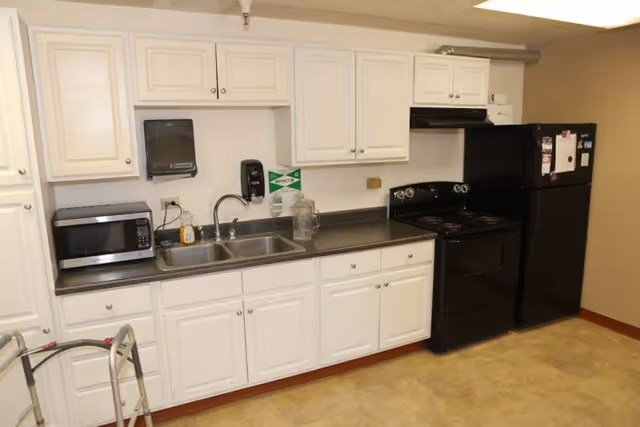 A kitchen area with white cabinets, a black stove, a black refrigerator, a microwave, a double sink, and a paper towel dispenser mounted on the wall. There is a walker partially visible in the foreground.