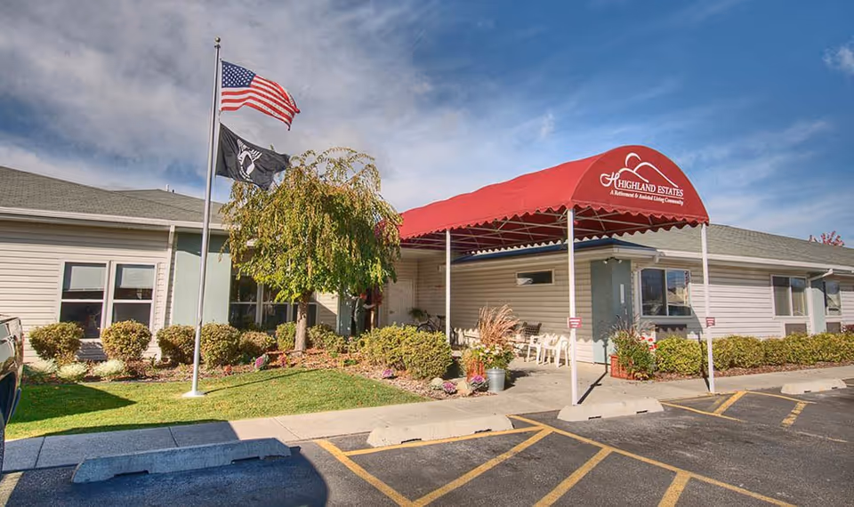 Front entrance of Highland Estates assisted living building with a red canopy, flagpoles, and landscaped shrubs.
