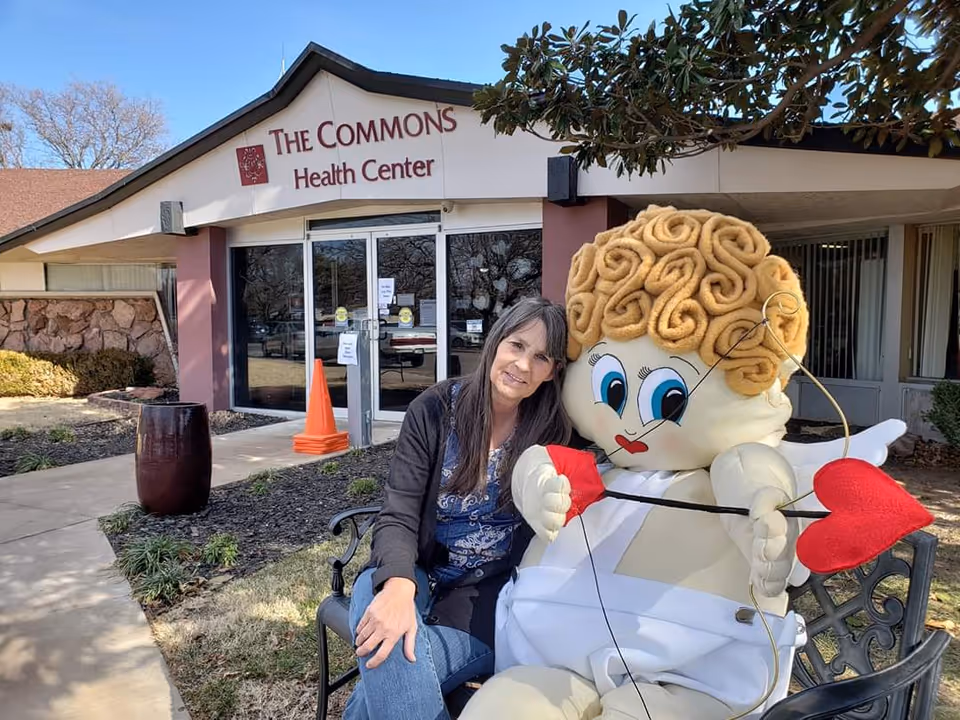 A woman sitting on a bench outside The Commons Health Center next to a large cupid mascot holding a bow with a heart-shaped arrow. The building entrance and some landscaping are visible in the background.