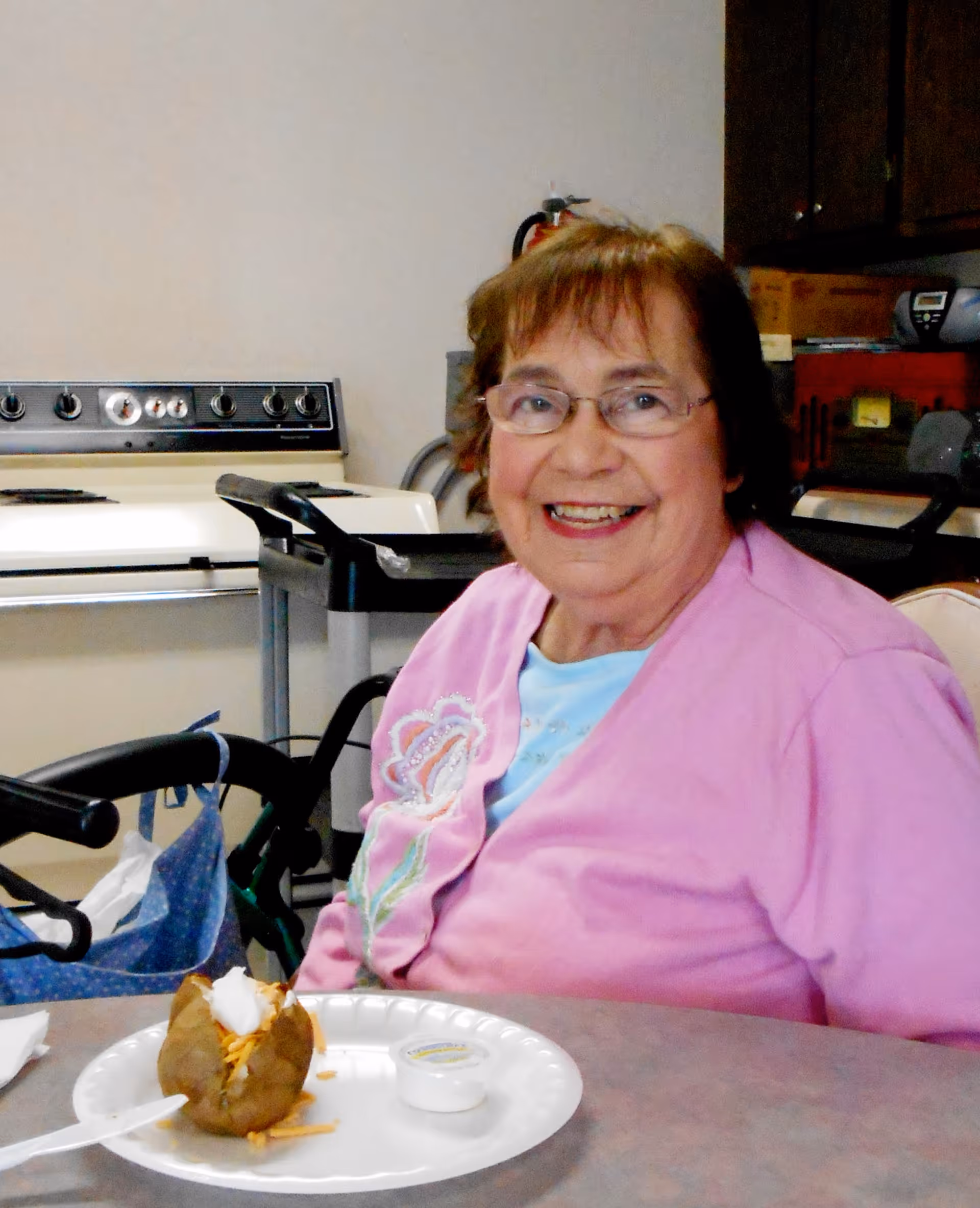 An elderly woman wearing glasses and a pink cardigan with floral embroidery is smiling while sitting at a table with a baked potato topped with sour cream and shredded cheese on a white disposable plate in front of her. Behind her is a kitchen area with a stove and cabinets.